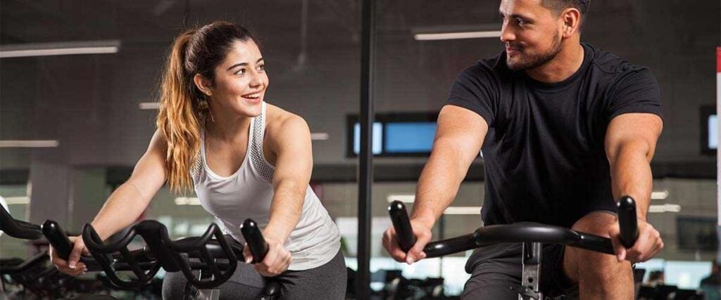 Man & Women Riding Spin Bikes At Gym