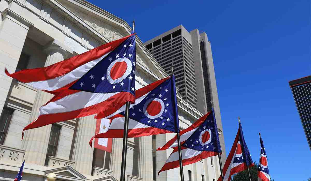 3 Ohio flags hung up on outside of building blowing in the wind on a sunny day.