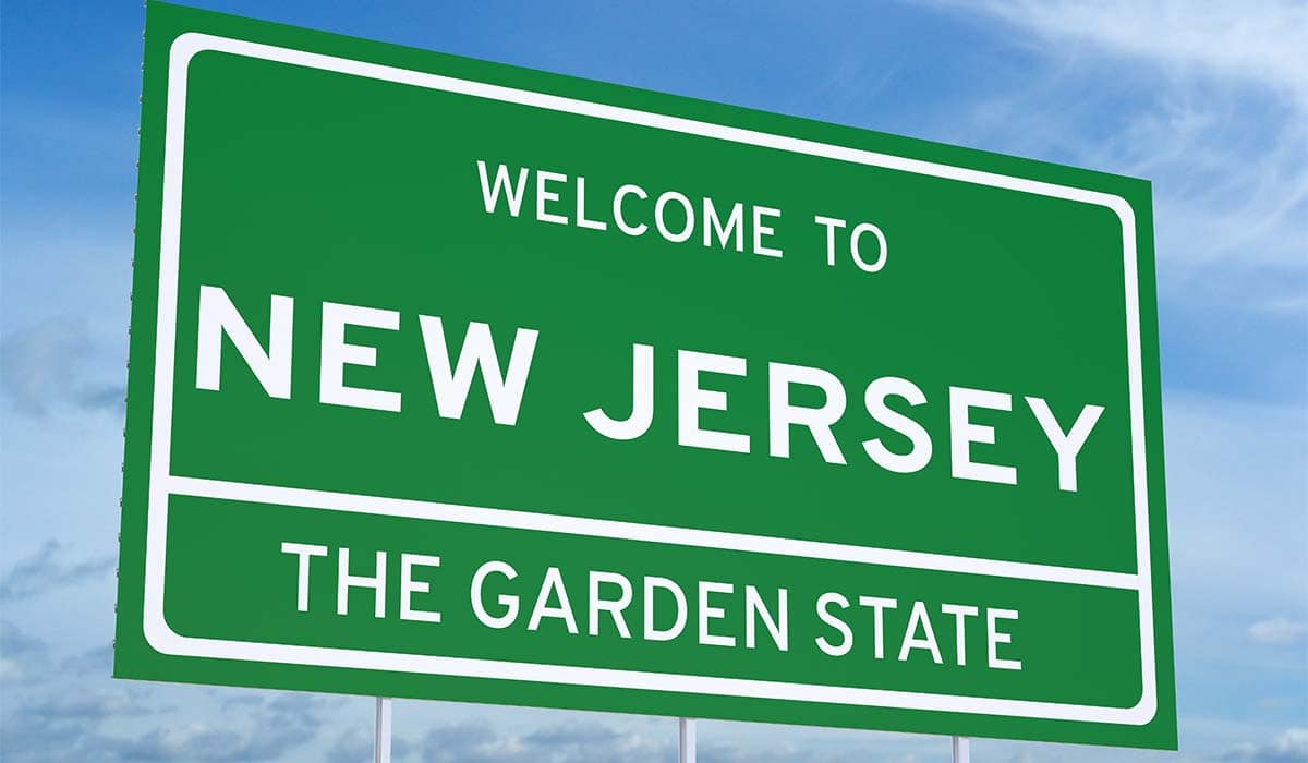 Large green 'Welcome to New Jersey: The Garden State' sign at the side of the road with blue sky as background. 