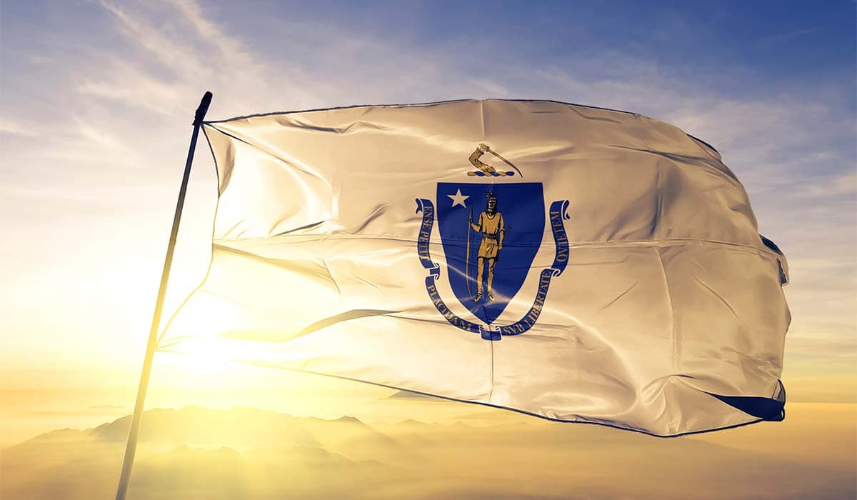 Massachusetts flag blowing in the wind with the sunset in the background and sun light pouring in from behind the flag.