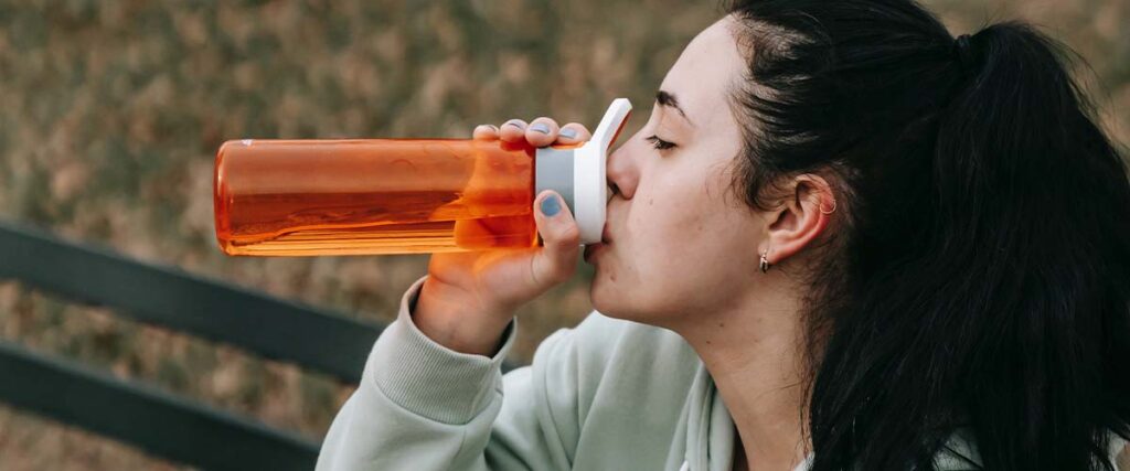 Woman drinking out of orange water bottle while on a park bench.