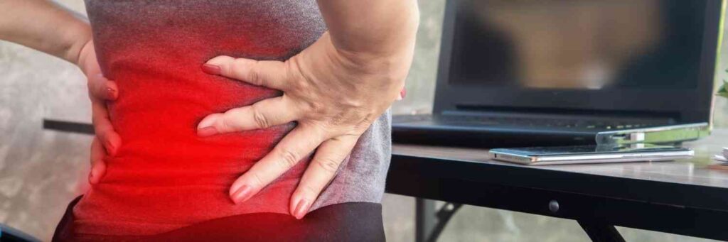 A woman on her computer holding her back with a red emphasis of where it hurts. 