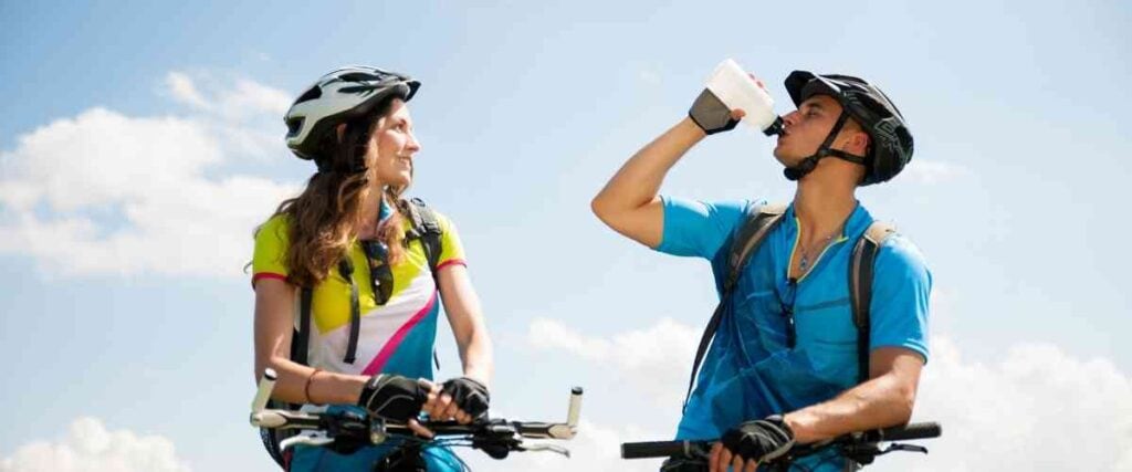 Two cyclists on the side of the road, one is drinking water. 