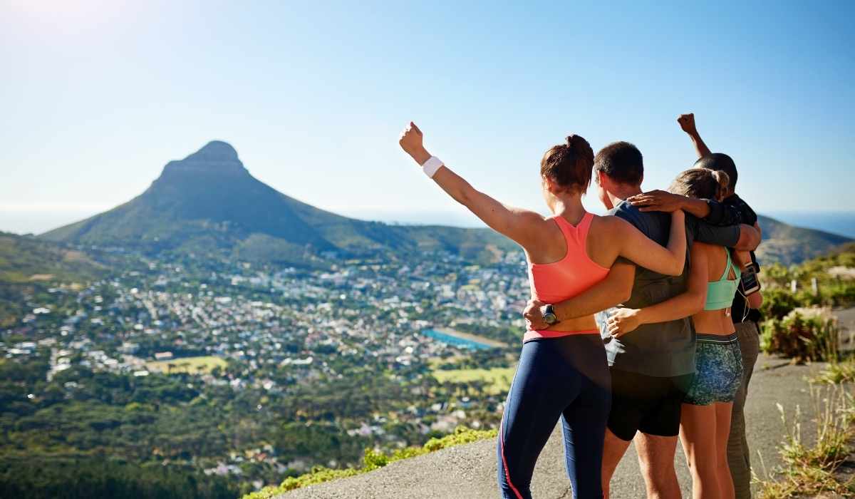 Group of men and women on top of a mountain, with their arms around each other celebrating while looking down over the city. 