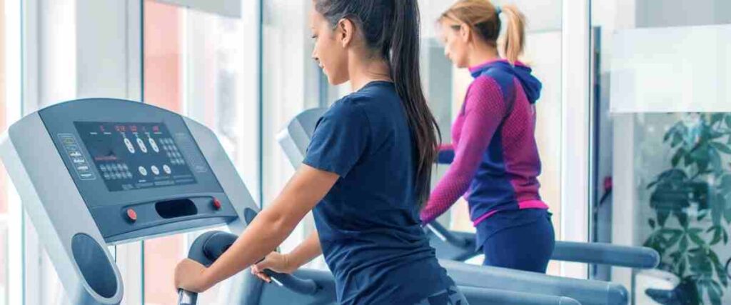 Two women adjusting the treadmill settings together on their own treadmill.