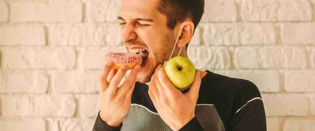 Man taking a bite out of a doughnut with an apple in the other hand. 