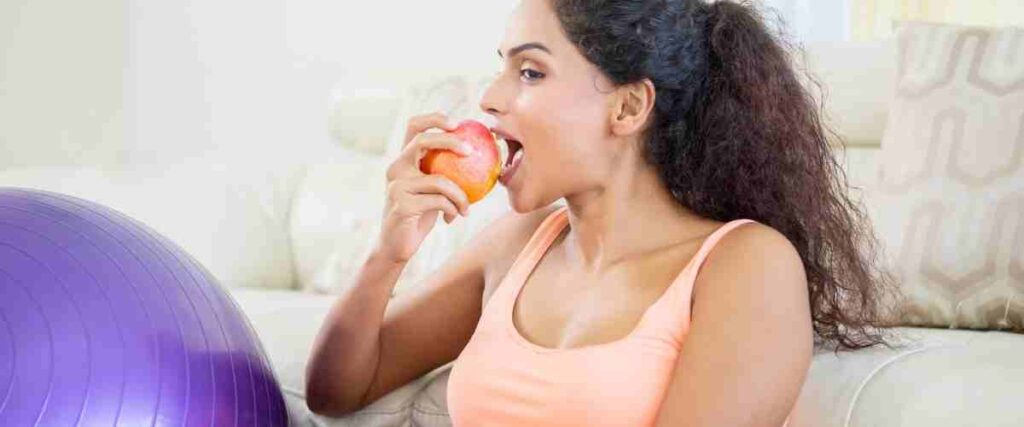 A woman eatting an apple next to her couch and workoutball. 