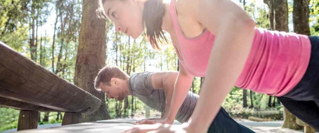 A couple doing push ups on a park bench in the woods. 