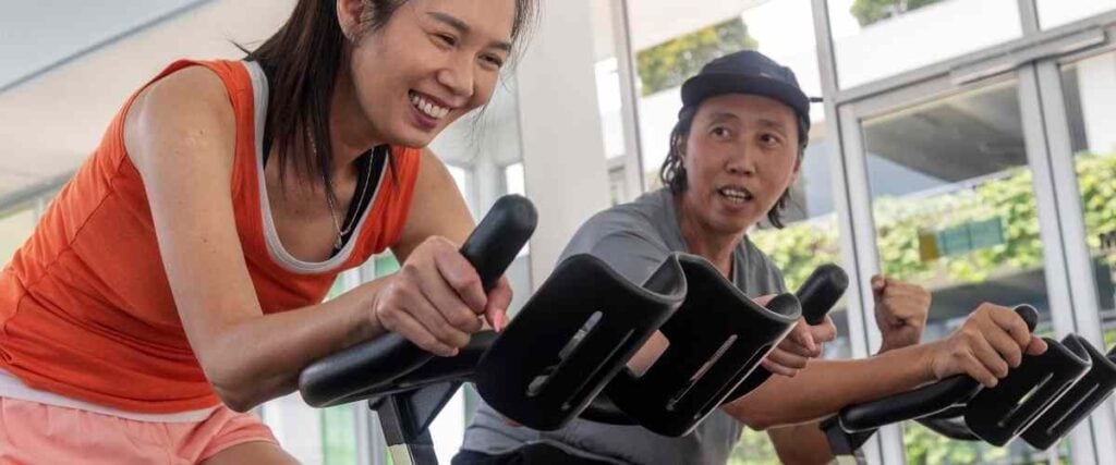 A couple on indoor exercise bikes at a gym next to a lot of windows.