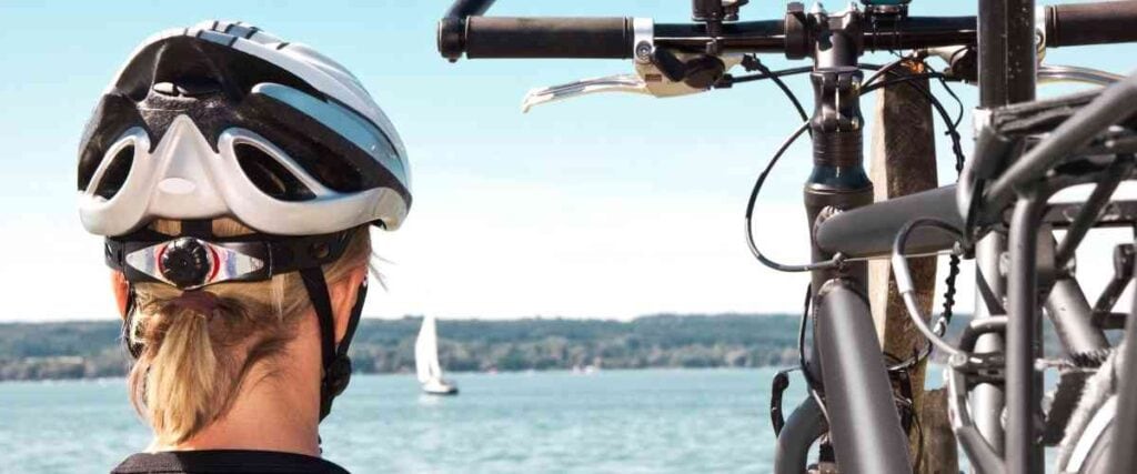 Woman with helmet on next to her bike, look at a sailboat on the sea. 