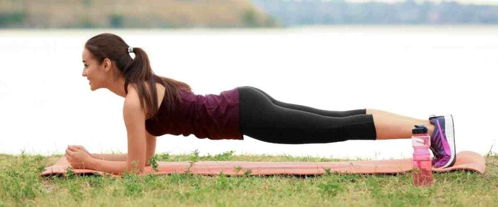 Woman doing the plank in the park on a yoga mat next to a lake. 