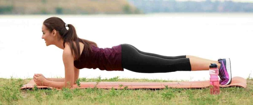 Woman doing the plank in the park on a yoga mat next to a lake.
