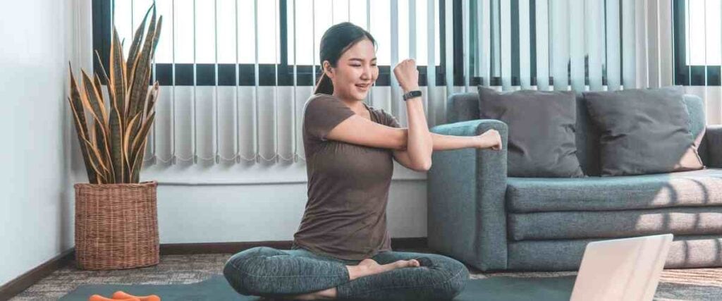 A woman doing stretches in her living room while looking at her computer.