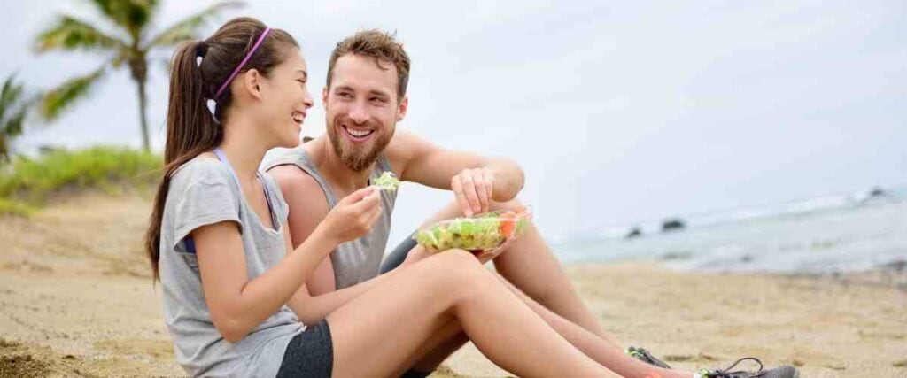 A couple laughing at the beach eating after their workout. 