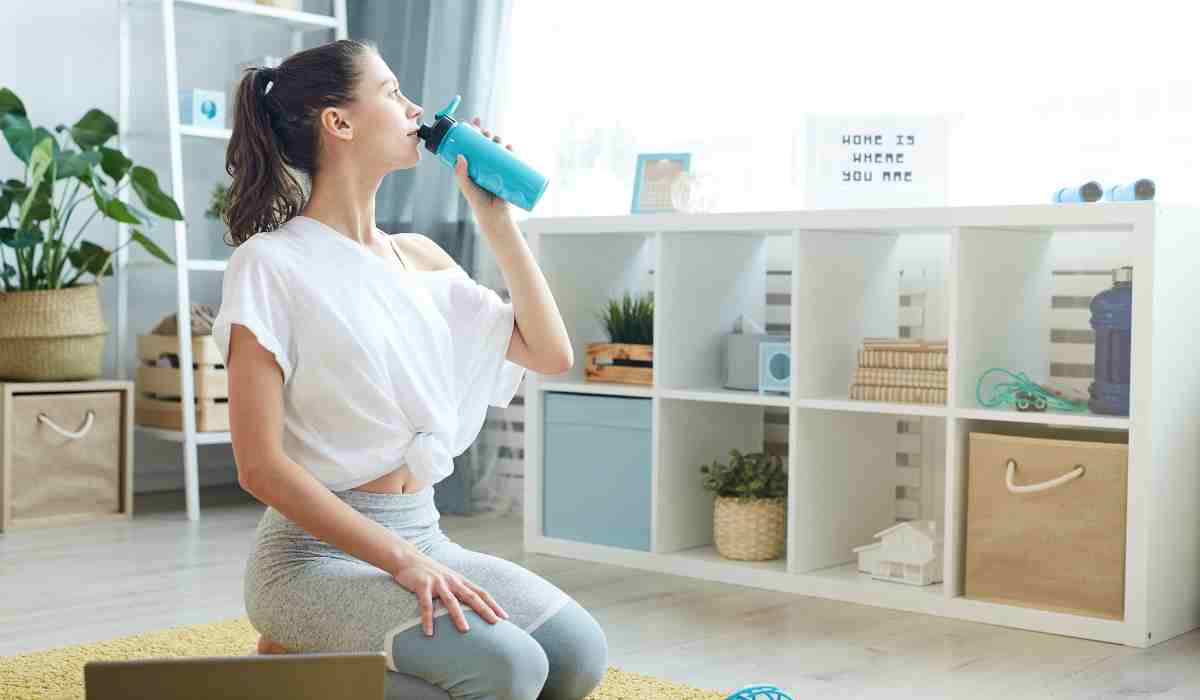 A woman drinking water after her workout on a yoga mat in her living room. 