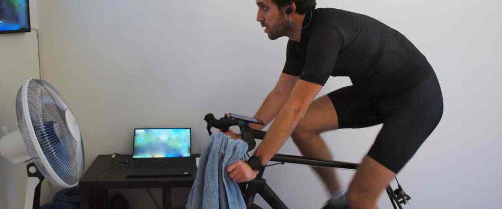 A male cyclists indoors working out while watching his tv with a fan blowing on him. 