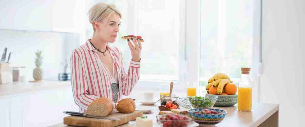 A woman in her kitchen surrounded by healthy foods while she is in mid-bite with toast in her mouth. 