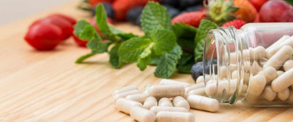 Supplements spilling out on a wooden surface with fruits and veggies behind the bottle of supplements.