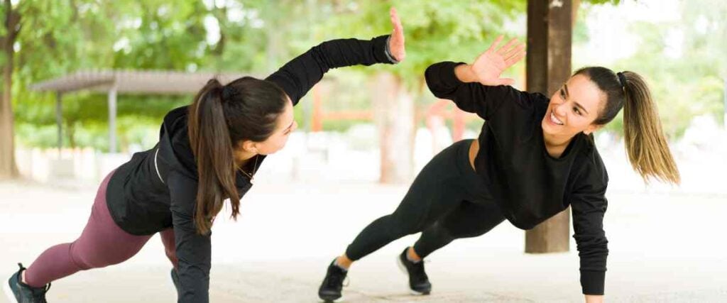 Two women high-fiving while doing a push up and smiling at each other at a park. 