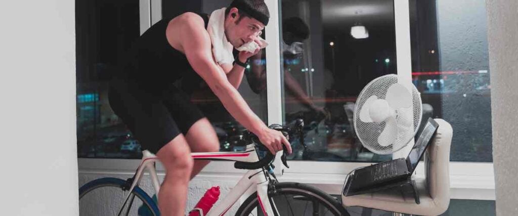 A male cyclists on his indoor trainer wiping his face with a towel, biking close to his laptop set up on a chair, fan next to big windows in his apartment. 