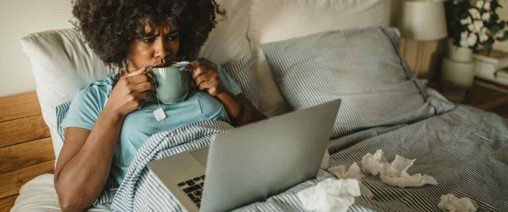 Woman sitting in her bed drinking tea sick, with used tissues in her bed since she is sick.