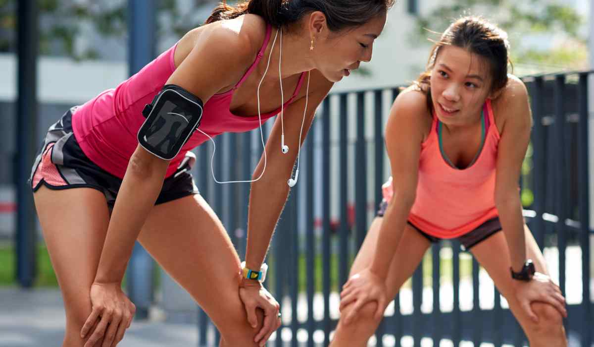 Two women hunched over trying to catch their breathe after a run next to an outdoor fence. 