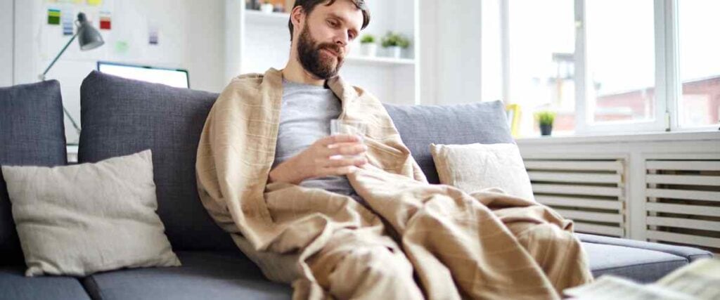 A man sitting on his couch wrapped in a blanket drinking water looking down at the floor.