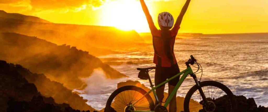Cyclists on top of the trail looking down on the ocean with her arms up - early in the morning watching the sun rise. 