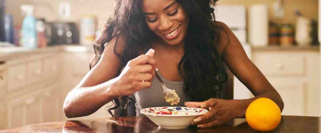 A woman eating oatmeal with an orange next to her while in her kitchen. 