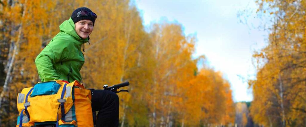 A man on his bike with additional bags during a bike ride during the fall with big trees covering the track with yellow leaves.