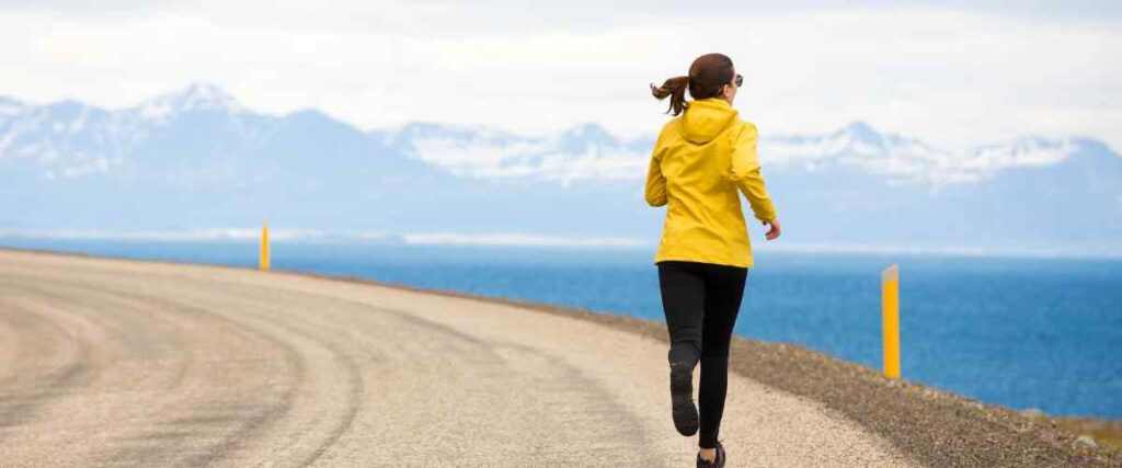 A woman running on a gravel road taking a turn with the ocean and mountains as the backdrop.
