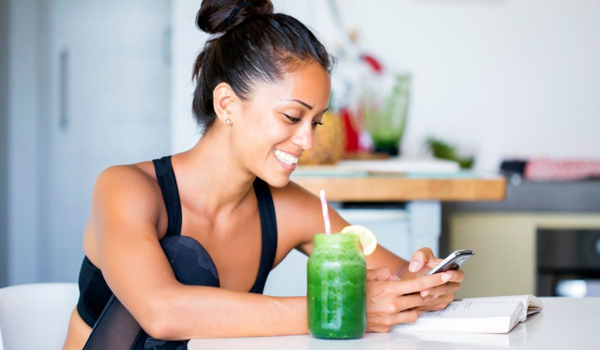 A woman on her phone after her workout drinking a smoothie. 
