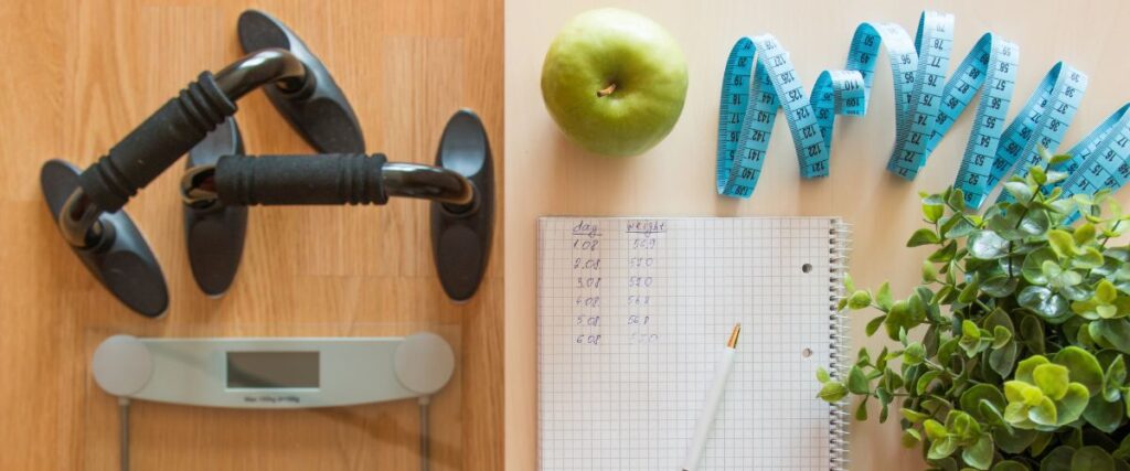 A scale and weights next to a notebook logging their weight loss journey. 