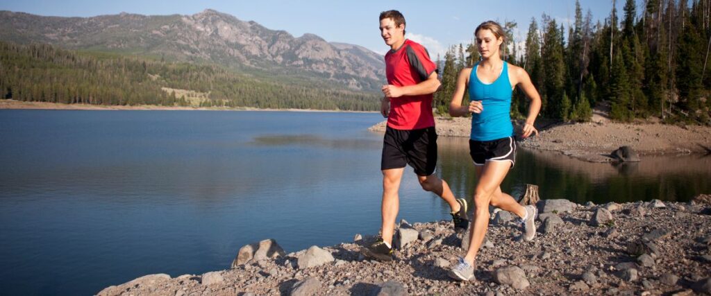 A couple running on a trail next to a stream and mountain. 