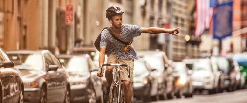 A young man cycling in NYC doing a right turn signal. 