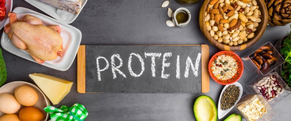 A sign with the word 'protein' on it with different types of protein around the sign like nuts, chicken, and rice. 