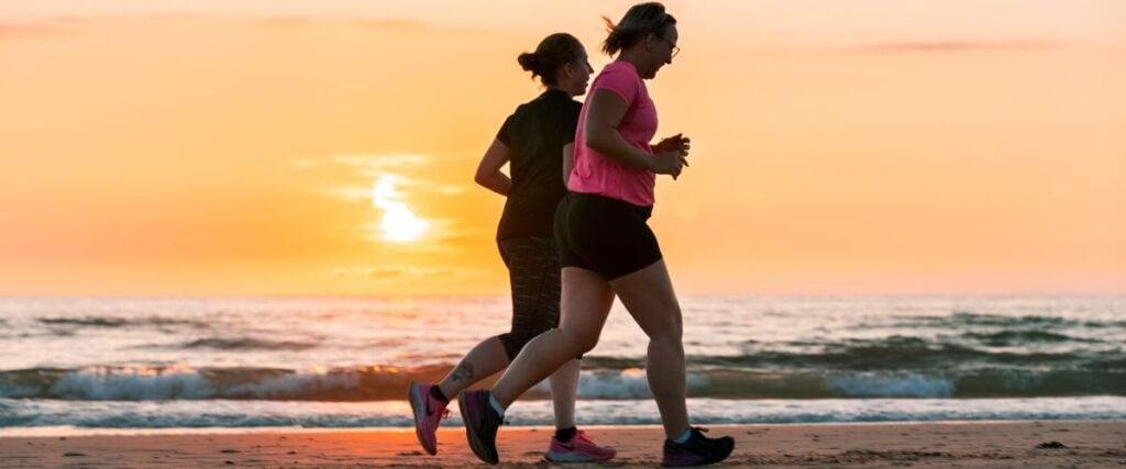 Two women jogging on a beach during sunset.