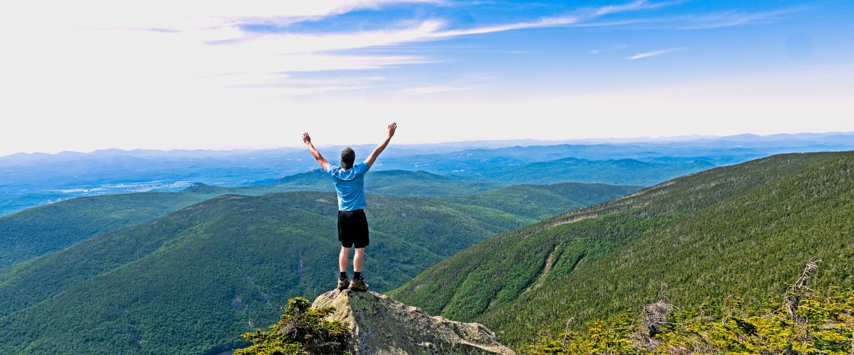 An accomplished hiker cheers on the top of a tall hill with a scenic view.