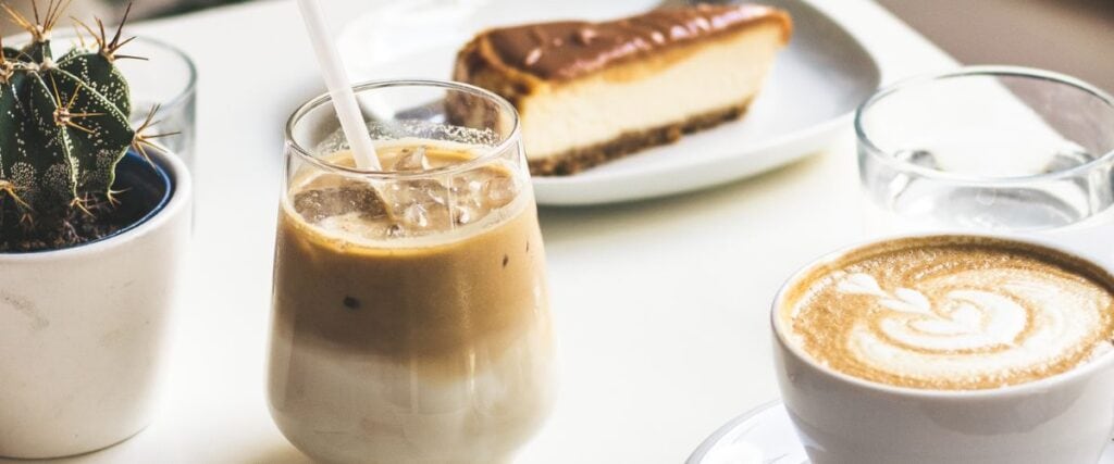 A cappuccino, iced coffee, and cheesecake are served on a table in a cafe.
